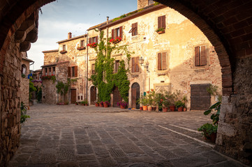Montemerano, Tuscany. View of the main square of the old medieval town through a red brick arch. Ivy stuck on the wall and ornamental plants. Walls illuminated by the light of the setting sun