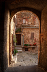 Fototapeta premium Montemerano, Grosseto, Tuscany. Glimpse of the ancient center of the medieval village seen through a stone arch. Stone floor and red brick walls