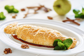 Baked Apple Strudel Cake Sweet Dessert Closeup. Classic Puff Pie with Vanilla and Mint Leaf on White Plate. Cinnamon with Walnut and Green Fruit on Blurred Background. Homemade Apfelstrudel Gourmet