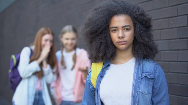 Sad bullying victim looking camera, female classmates teasing african schoolgirl