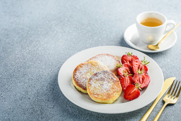 Cottage cheese pancakes or Syrniki with fresh strawberries on a white plate. Curd fritters with a cup of tea for breakfast
