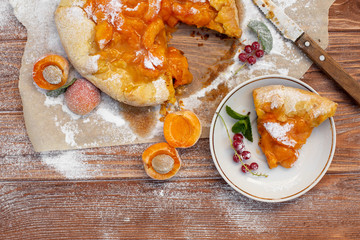 Homemade fruit pie (galette) made with fresh organic apricotes with powdered sugar on wooden table, top view. Plate with a cut piece of cake. Open pie, apricot tart. Summer pie on parchment paper.