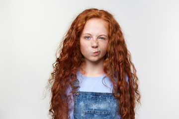 Photo of malcontent nice freckles little girl with ginger hair, parents forbid to watch cartoons, grumpy looks at thecamera, stands over white background.
