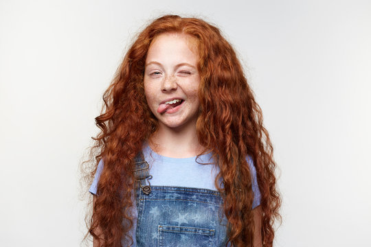 Portrait of funny cute freckles little girl with ginger hair, winks and shows tongue at the camera, stands over white background.