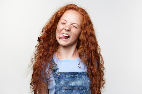 Close up of joyful cute little girl with ginger hair and freckles, shows tongue at the camera and looks funny, stands over white background with closed eyes.