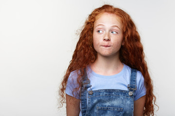 Portrait of curious cute freckles little girl with ginger hair, thinking about something, bites...