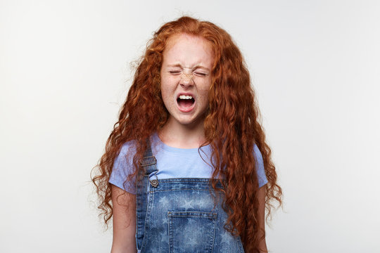 Prtrait of sceaming freckles little girl with ginger hair, stands over white wall with closed eyes and wide open mouth, looks unhappy and crying.