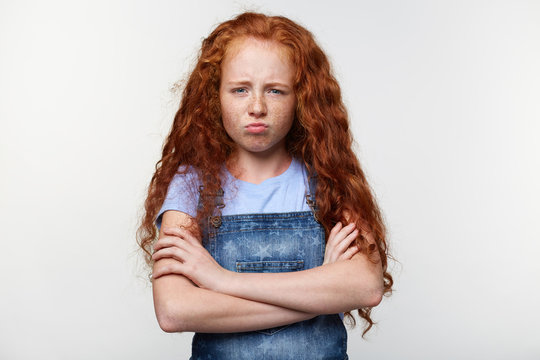 Prtrait of unhappy freckles little girl with ginger hair, stands over white wall with crossed arms, looks sad and offended.