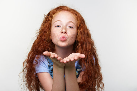 Portrait of cheerful little girl with ginger hair and freckles, send kiss at the cam, looks happy, standing over white wall.