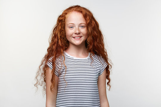 Portrait of positive cute freckles little girl with ginger hair, stands over white wall and broadly smiling, looks happy and glad.