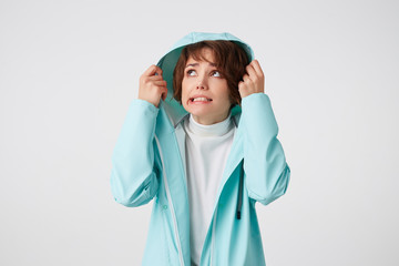 Cute disgusting short-haired curly woman in white golf and light blue rain coat, hiding under the hood from rain and looking up at the left side, stands over white background.