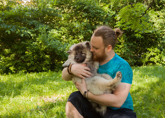 Young man with a red beard in a turquoise T-shirt is holding a Keeshond puppy.  Early summer evening.