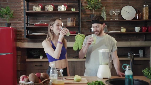Slow motion close up view of beautiful young Caucasian man and woman in sportswear raising glasses with organic fresh smoothie, drinking it with pleasure and joyfully dancing. Positive emotions