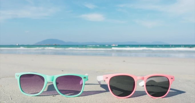 On a tropical beach close-up sunglasses pink and blue lie on the white sand against the ocean.