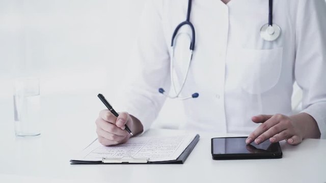Female caucasian doctor using tablet and writing diagnosis in medical form and documents. Close up 4K