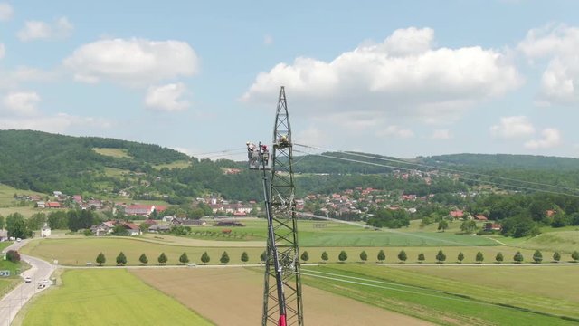 AERIAL: Flying around electricians standing inside a telescopic boom lift and repairing an electricity pylon. Maintenance crew servicing high voltage electricity cables on a tall metal power tower.