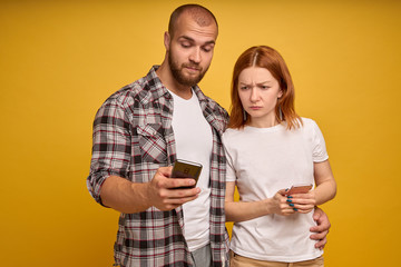 Portrait of a young couple standing with mobile phone, man is using mobile phone while angry girl standing near isolated over yellow background
