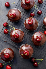 Chocolate-coffee muffins with melted dark chocolate topping with the addition of fresh cherries on a cooling tray on a dark background, top view. Delicious dessert