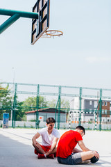Two strong caucasian athletes resting on the ground at the basketball court while having a conversation.