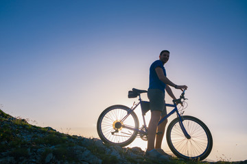 Obraz na płótnie Canvas Strong fit male mountain biker performing stunts on rocky terrain on a sunset while wearing a blue shirt and riding a blue bike