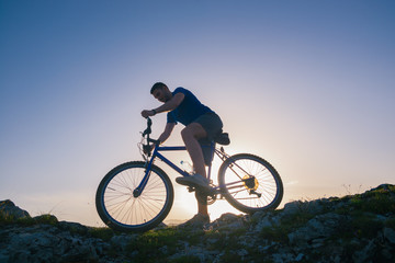 Obraz na płótnie Canvas Strong fit male mountain biker performing stunts on rocky terrain on a sunset while wearing a blue shirt and riding a blue bike