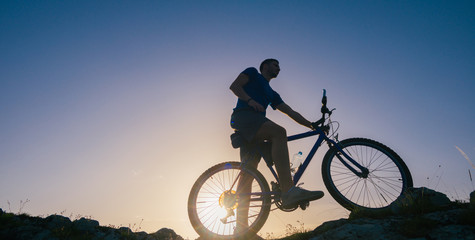 Fototapeta na wymiar Silhouette of a fit male mountain biker riding his bike uphill on rocky harsh terrain on a sunset.