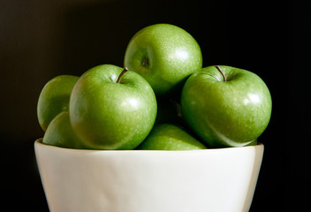 A group of green Granny Smith apples in a white fruit bowl against a dark brown background.