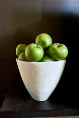 A white bowl full of green Granny Smith apples against a brown wall and table.