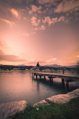 Lonely man standing on the edge of the wooden pier looking at the calm lake and forest on the other...