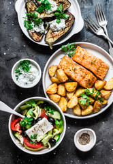 Greek style lunch table - baked lemon salmon with potatoes, greek salad, grilled eggplant with tzadziki sauce on dark background, top view. Flat lay
