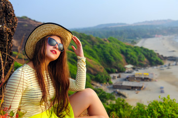Portrait of an attractive young girl with long hair in straw hat and sunglasses on background of green hills. Selective focus. Beautiful landscape, rest, tourism, travel.