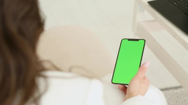 girl brunette in a white jacket looks into the phone leafs through pictures, green background, chromakey, looks out the window, near the table with a laptop