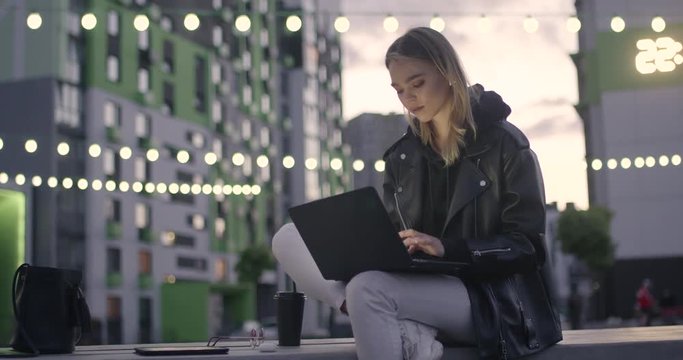 Young girl in wireless headphones uses a laptop sitting outside in a modern residential area, evening in the city, blurry lights on background, 4k slow motion.