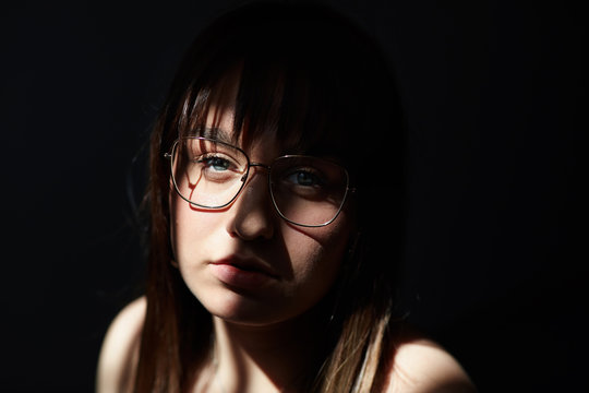 Close up sensual portrait of young blue eyed Caucasian female with dark hair and charming appearance, sitting in dark room under sunny ray, wearing big elegant eye glasses, looking at camera deeply.