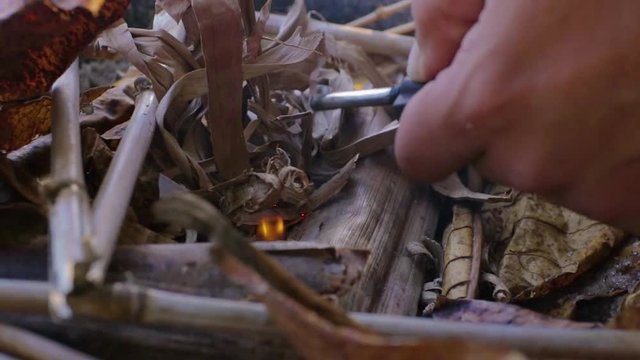 Close up of a man trying to start a fire with a  some metal knife, during a sunny day, in a camping ate the beach, just the hands can be seen in the shot