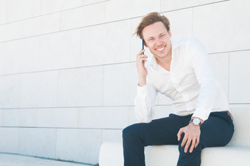 Happy business man talking on mobile phone on bench outdoors. Guy looking at camera and sitting on stone bench at building wall. Communication in business concept. Front view.
