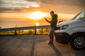 Young handsome man use phone standing near his car on beautiful sunset on sea background, Travel, summer vocation.