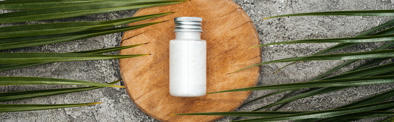 top view of coconut beauty product in bottle on wooden board near palm leaves on grey textured background, panoramic shot