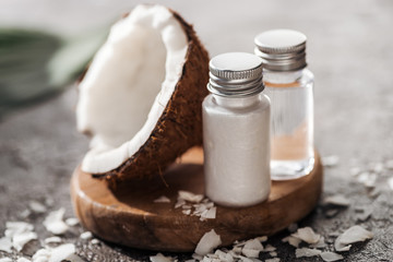 selective focus of bottles with coconut beauty products on wooden board near coconut half on grey textured background