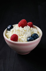pink bowl with cottage cheese, blueberries and raspberries on a black wooden background