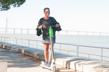 Happy joyous black woman enjoying riding scooter. Cheerful Afro American female rider posing on scooter near river. Scooter rider concept