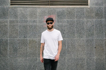 Hipster handsome male model with beard wearing white blank t-shirt and a baseball cap with space...