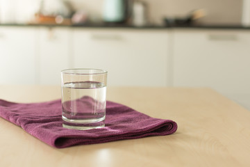 Glass of pure water on kitchen table on purple napkin. Fresh transparent water in a bright modern kitchen blur background. Copy space for design.