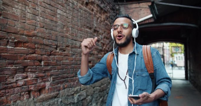 Happy Arab tourist in headphones listening to music singing dancing outdoors having fun alone in the street holding smartphone. Youth culture and devices concept.