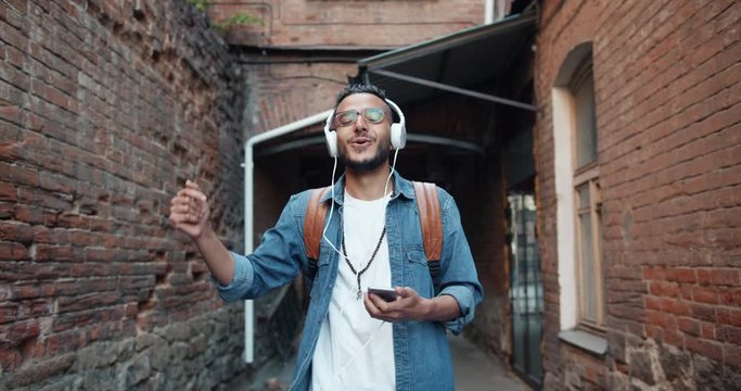 Slow motion of African American guy in headphones dancing singing outdoors holding smartphone having fun in the street. People, youth and devices concept.