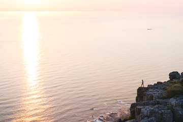 A single person on a cliff above the ocean,silhouetted against the reflection of the setting sun. - 276884983