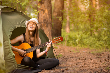 Fototapeta na wymiar Tourist girl in hat sits in tent and playing guitar concept. Tourism rest on nature. Camping lifestyle