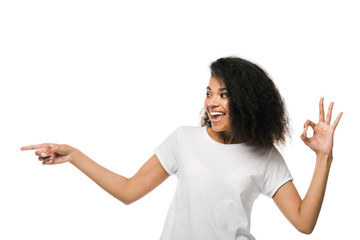 cheerful african american woman pointing with finger while showing ok sign isolated on white