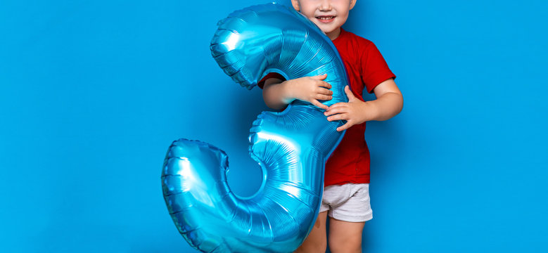 small cute blonde boy on blue background holding foil-coated sphere baloon blue colour. happy birthday three years old