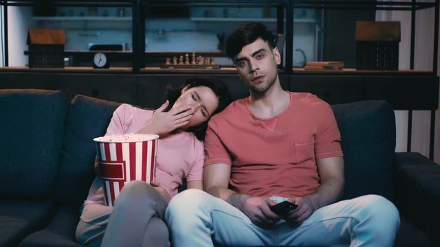 young woman holding paper cup with popcorn and yawning while leaning of shoulder of man holding remote controller and watching tv at home